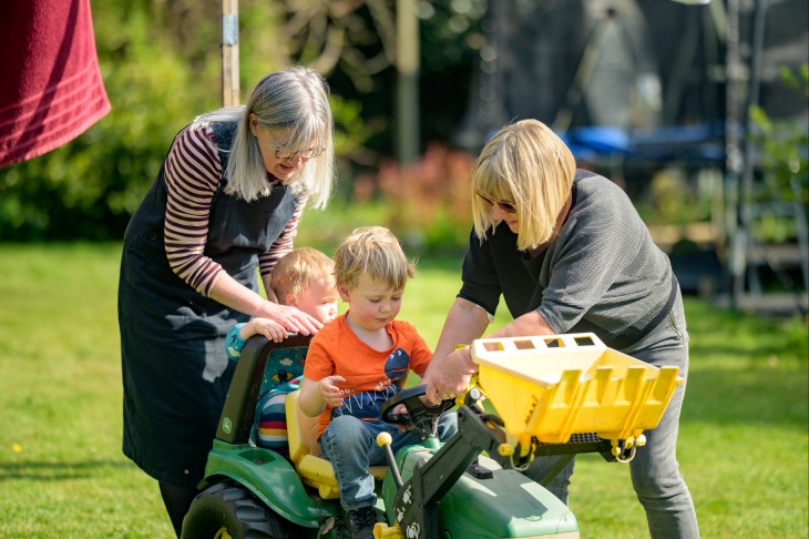 Two adult women help two young children playing on a toy tractor