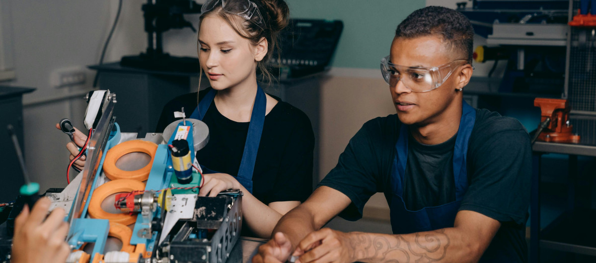 Two young people in a factory, being taught to work a specific tool